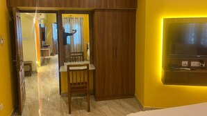 A modern room with a wooden wardrobe, a small table with a chair, a TV mounted on the wall, and a bathroom visible through a glass door. - White Yellow Residency (Puducherry)