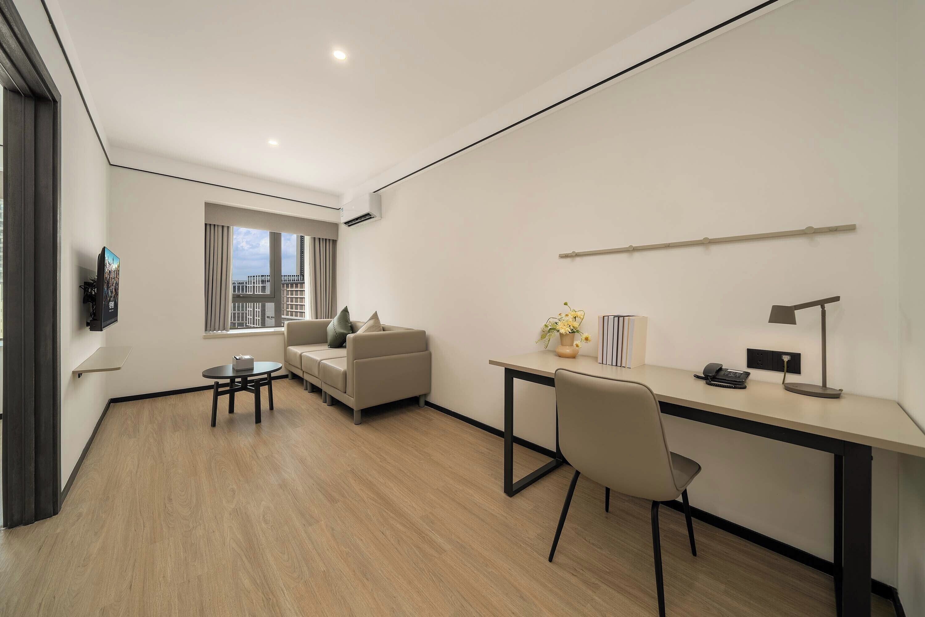 Habitación moderna y bien iluminada con sofá, una pequeña mesa redonda, un escritorio con silla, televisor montado en la pared y ventana con vistas a la ciudad.