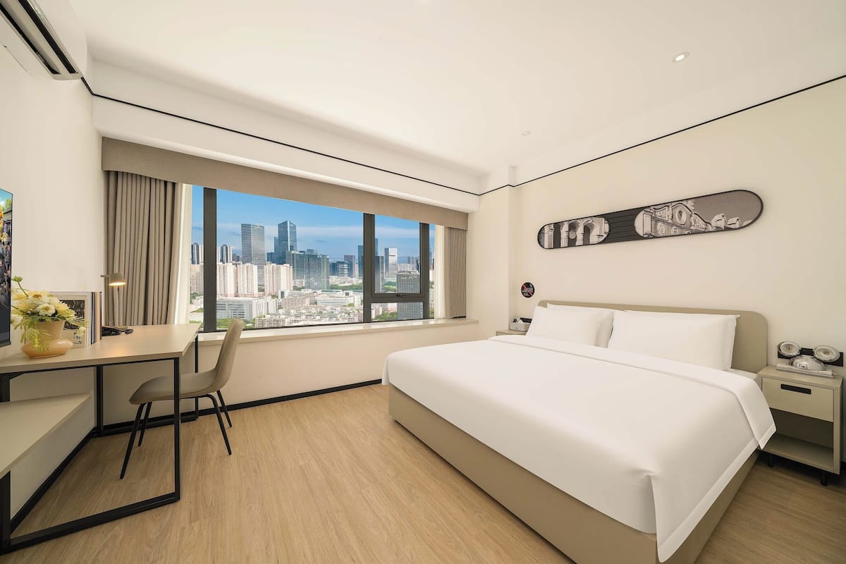 Ein Hotelzimmer mit einem großen Bett, einem Schreibtisch und einem Stuhl. Ein Fenster bietet Blick auf die Stadt.