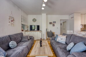 Una sala moderna con dos sofás grises, una mesa de centro de madera, un televisor de pantalla plana y una zona de cocina al fondo.