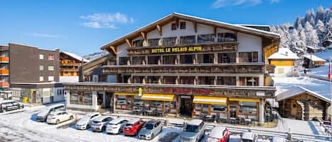 Un hôtel de style architectural alpin traditionnel, doté d’un balcon imposant et d’une enseigne indiquant « HÔTEL LE RELAIS ALPIN ».