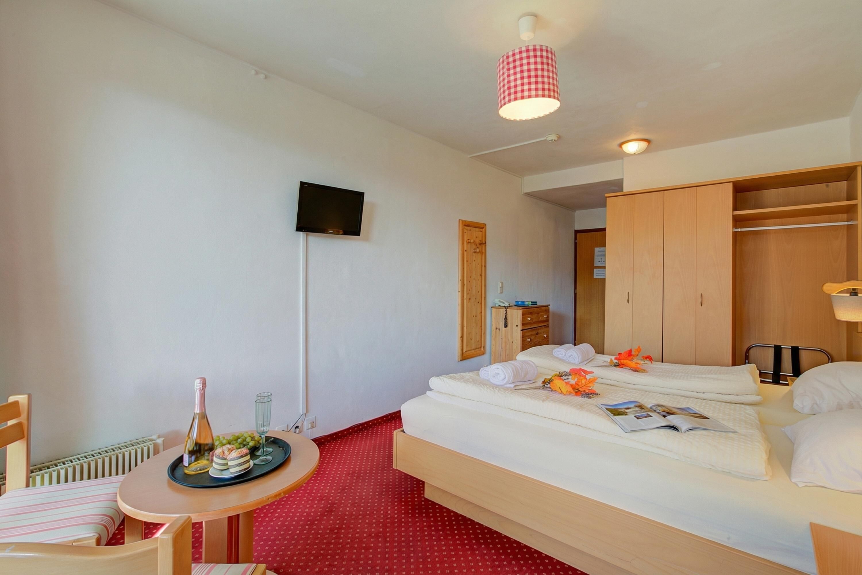 A hotel room with a bed, a small table with a bottle and glasses, a TV on the wall, and a wardrobe.