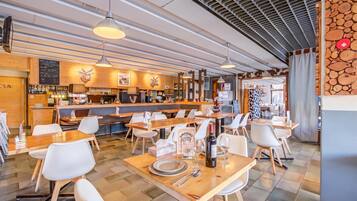 Ein Restaurantinneres mit Holztischen, weißen Stühlen, hängenden Pendelleuchten und einer Bar mit einer Speisekarte.
