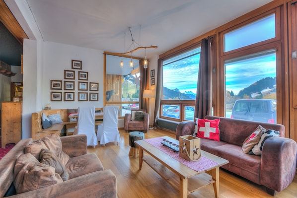 Ein gemütliches Wohnzimmer mit einer Couch, einem Tisch und Blick auf Berge durch große Fenster.