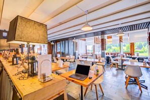 Un restaurant moderne avec des tables et des chaises en bois, un coin bar équipé d’un ordinateur portable et d’une machine à café, et de grandes fenêtres donnant sur l’extérieur.