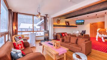 Ein geräumiges Wohnzimmer mit Kamin, bequemen Sitzgelegenheiten, einem Fernseher und einem großen Fenster mit Blick auf eine verschneite Landschaft.