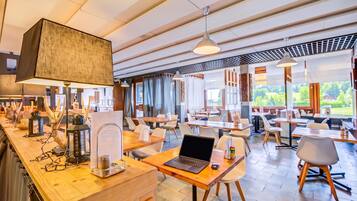 Ein modernes Restaurant mit Holztischen und -stühlen, einem Barbereich mit einem Laptop und einer Kaffeemaschine sowie großen Fenstern mit Blick ins Freie.