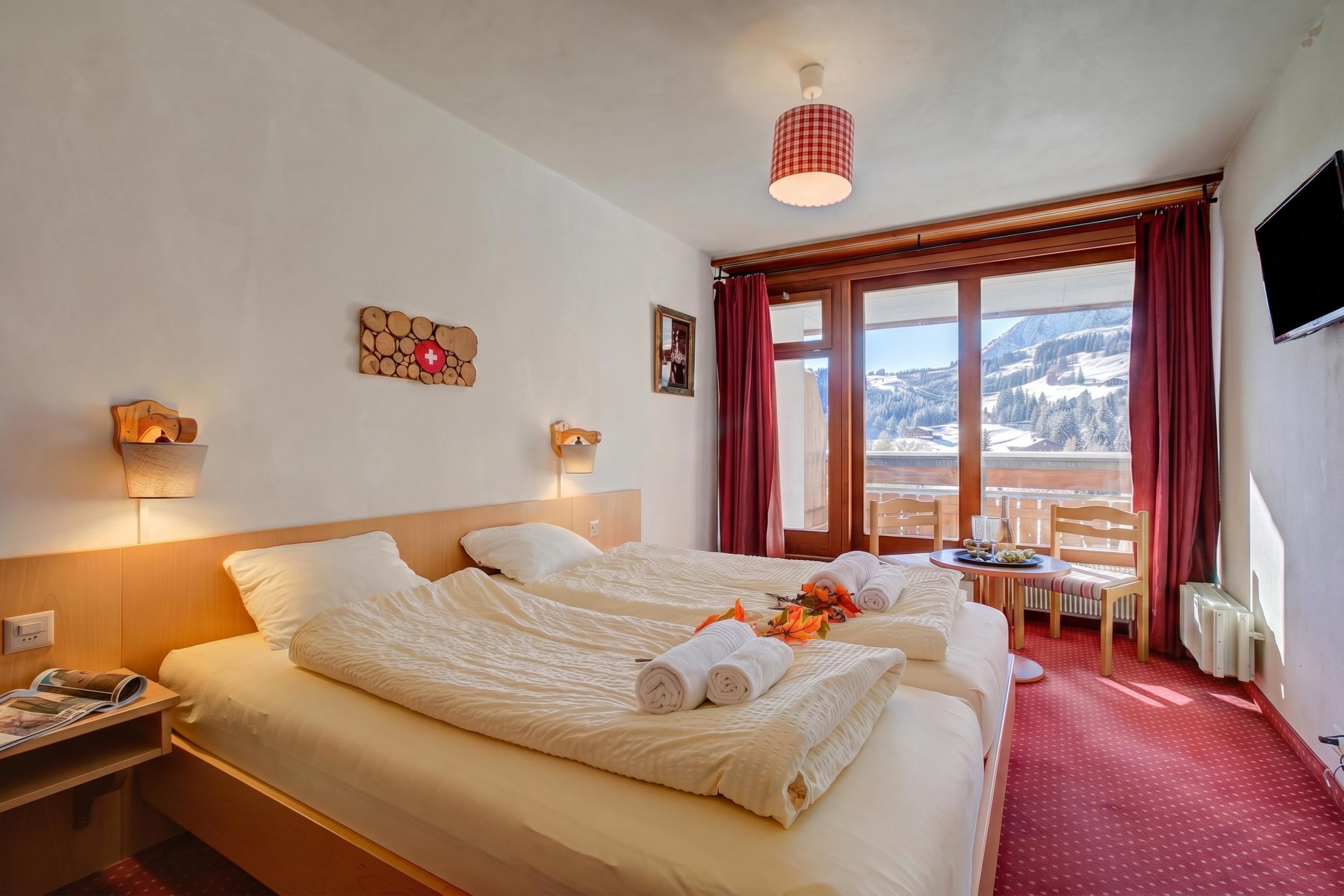 Habitación de hotel con cama, escritorio, silla, mesa con botella y vasos, televisión y ventana con vista a la montaña.