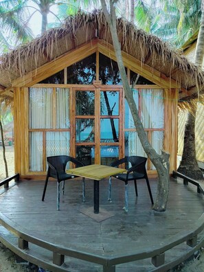A wooden hut with a thatched roof, large windows, and a wooden deck with two chairs and a table. - Aroma Beach Resort Saintmartin (Saint Martin's Island)