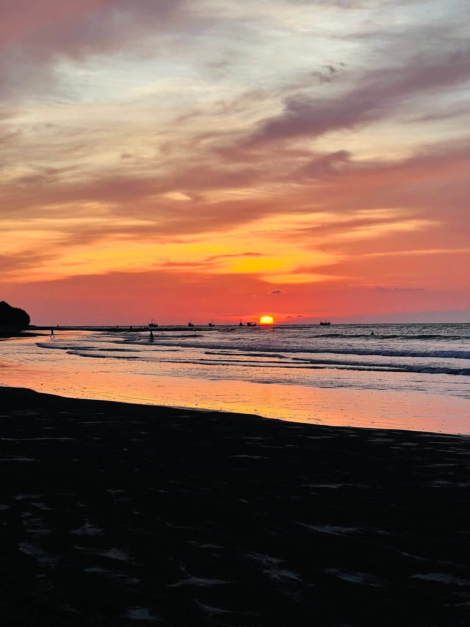 Cena de praia com um pôr do sol, mar calmo e barcos ao longe.