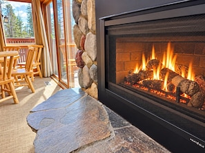 A stone fireplace with a fire burning inside, surrounded by a stone wall and wooden chairs.