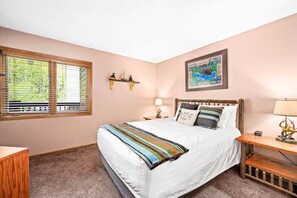 A bedroom with a large bed, a nightstand, a wall-mounted shelf, a window with blinds, and a painting on the wall.
