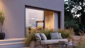 Ein moderner Sitzbereich im Freien mit einer Couch und einem Tisch, umgeben von hohem Gras und einem großen Fenster mit Blick auf einen Essbereich.