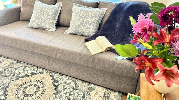 A cozy living room with a grey sofa, floral cushions, a blue throw blanket, and a book. A vibrant flower arrangement is on a side table. - Private Entire Guest Suite, 1 min to bus stops to Downtown and many attractions. (Vancouver)