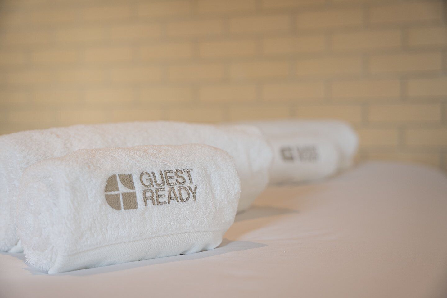 Dues tovelles blanques amb la frase "GUEST READY" brodada, col·locades sobre un llit, amb un mur de maó al fons.