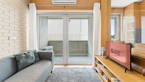 Een moderne woonkamer met een grijze bank, een flatscreen televisie op een houten standaard en een schuifdeur van glas die leidt naar een buitenbalkon.