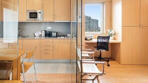 Een moderne keuken met houten kastjes, een magnetron, een waterkoker en een broodrooster. Er is een glazen scheiding die de keuken van een werkruimte met een bureau en bureaustoel scheidt.