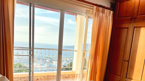 A hotel room with a large bed, a view of the city through a balcony, and wooden furniture. - Sunset Ocean View 3-king bed Villa Directly Above Patong (Phuket)