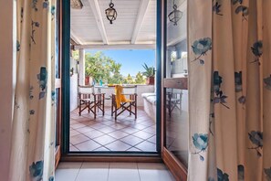 Vista através de uma porta de vidro para uma área de pátio externa com mesa, cadeiras e plantas em vasos.