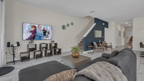 A modern living room with a large flat-screen TV, a staircase, and a dining area. - Three Themed Worlds. One Unforgettable Family Stay. (Kissimmee)