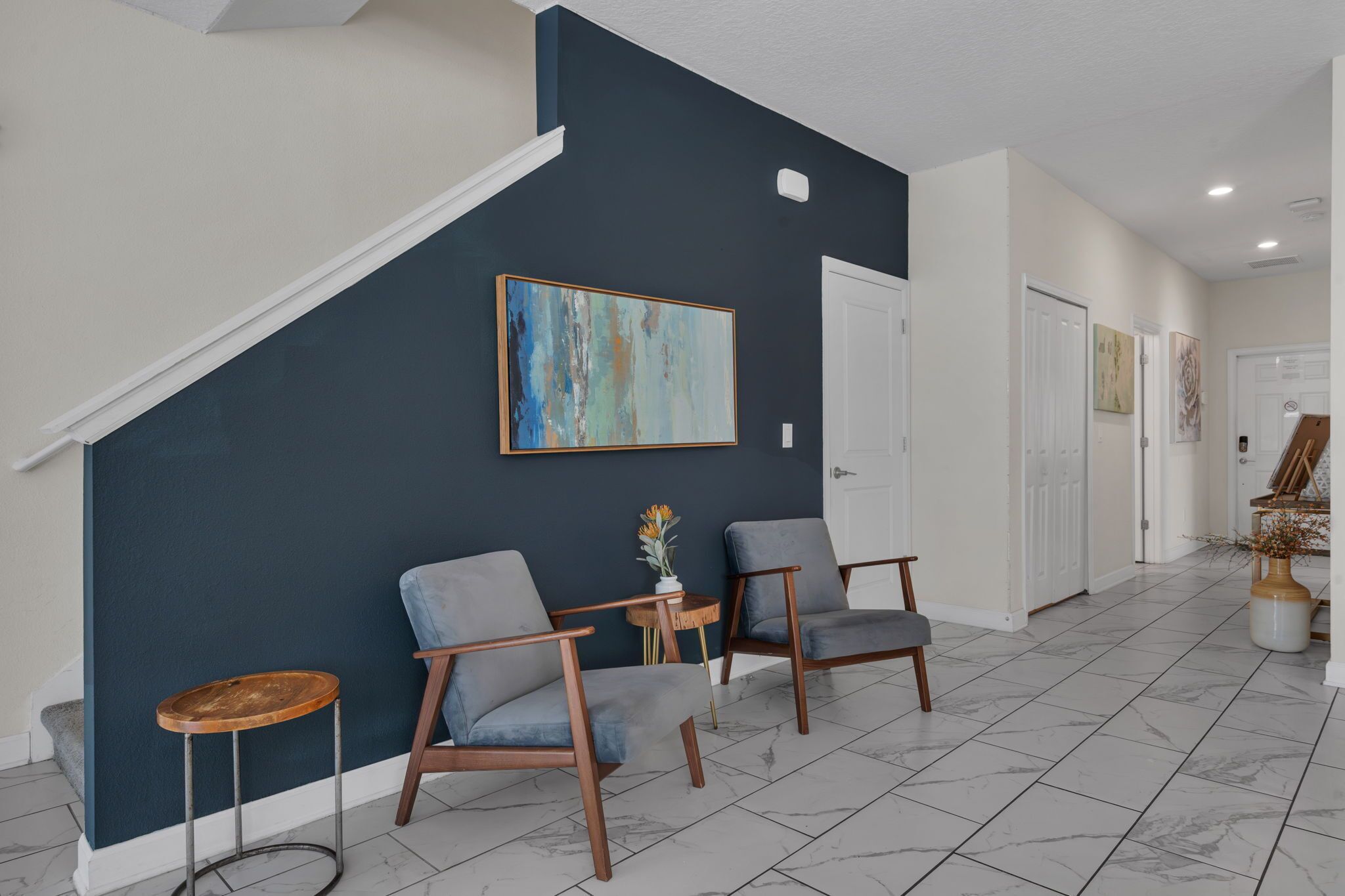 A modern interior with a dark blue accent wall, two grey armchairs, a small wooden table with a vase, and a staircase leading to another level.