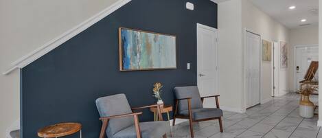 A modern interior with a dark blue accent wall, two grey armchairs, a small wooden table with a vase, and a staircase leading to another level.