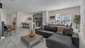 Uma sala de estar moderna com um sofá cinzento, uma mesa de centro e uma área de jantar com cozinha ao fundo.
