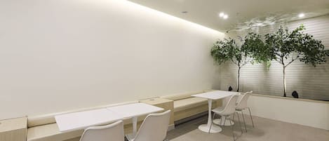 Un área de espera moderna con mesas y sillas blancas, un banco y una planta en la pared.