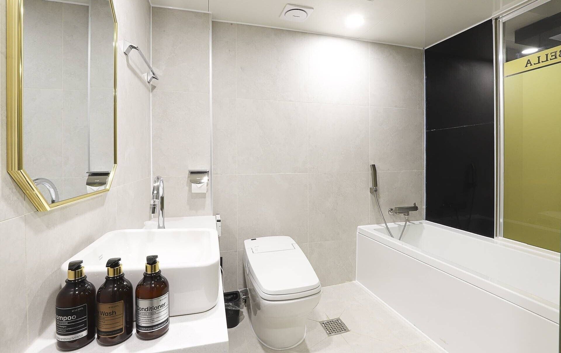 Un baño moderno con lavamanos, inodoro y bañera blancos. Hay tres botellas de champú, acondicionador y gel de baño sobre el mostrador.