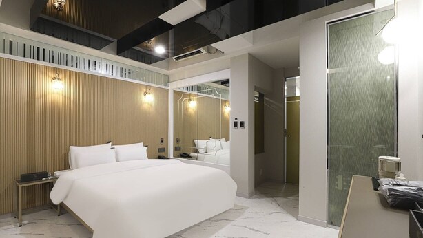 A modern hotel room with a large bed, a wooden panel wall, a mirror, and a bathroom with a glass door. - Swissbella Hotel (Pohang)