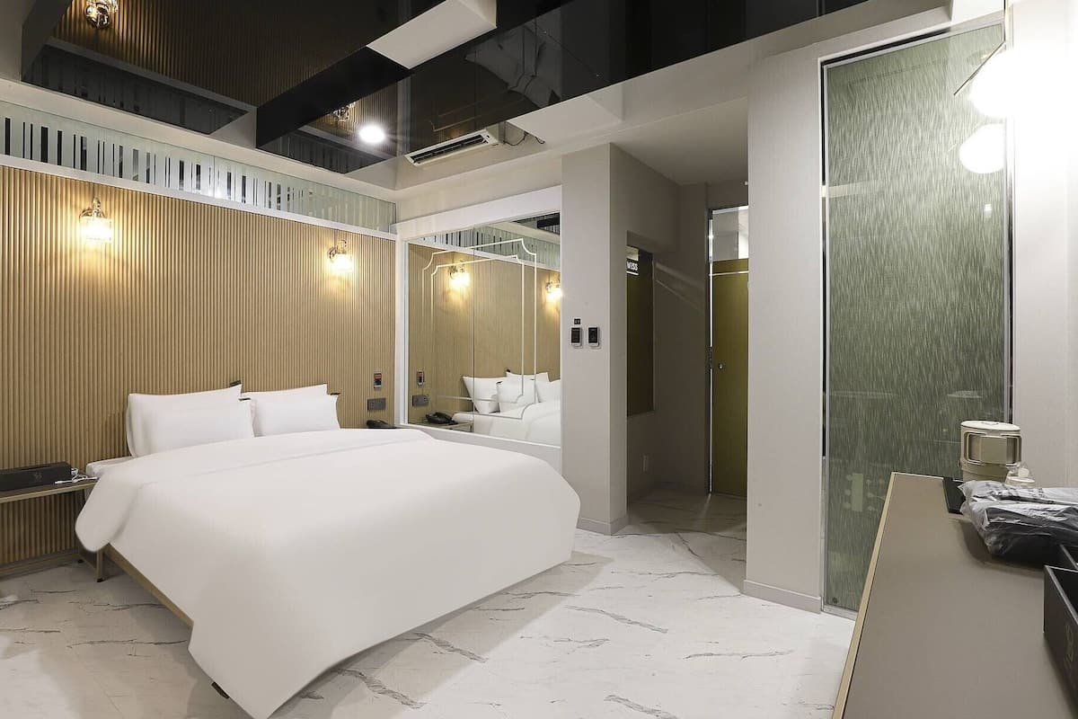 A modern hotel room with a large bed, a wooden panel wall, a mirror, and a bathroom with a glass door.