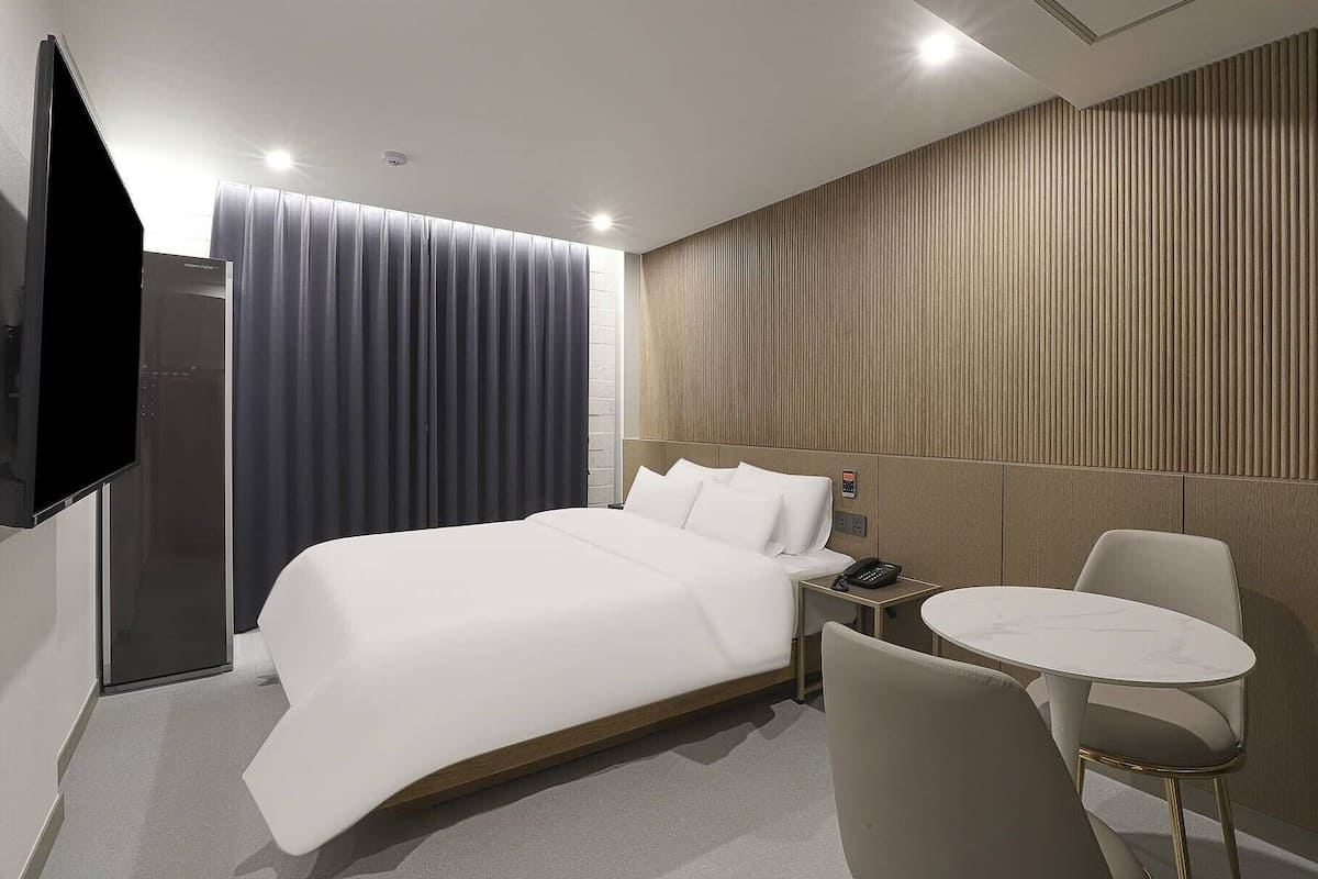 A modern hotel room with a large bed, a bedside table with a phone, a small round table, and a chair.