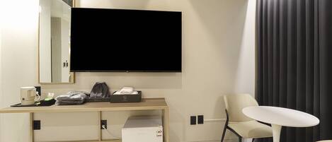 Una habitación de hotel moderna con televisor de pantalla plana en la pared, un escritorio con cafetera, un refrigerador pequeño y una silla.