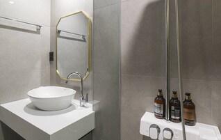 Un baño moderno con un lavamanos blanco, un espejo con marco dorado y una ducha con manguera. Hay tres botellas de artículos de tocador en la pared.