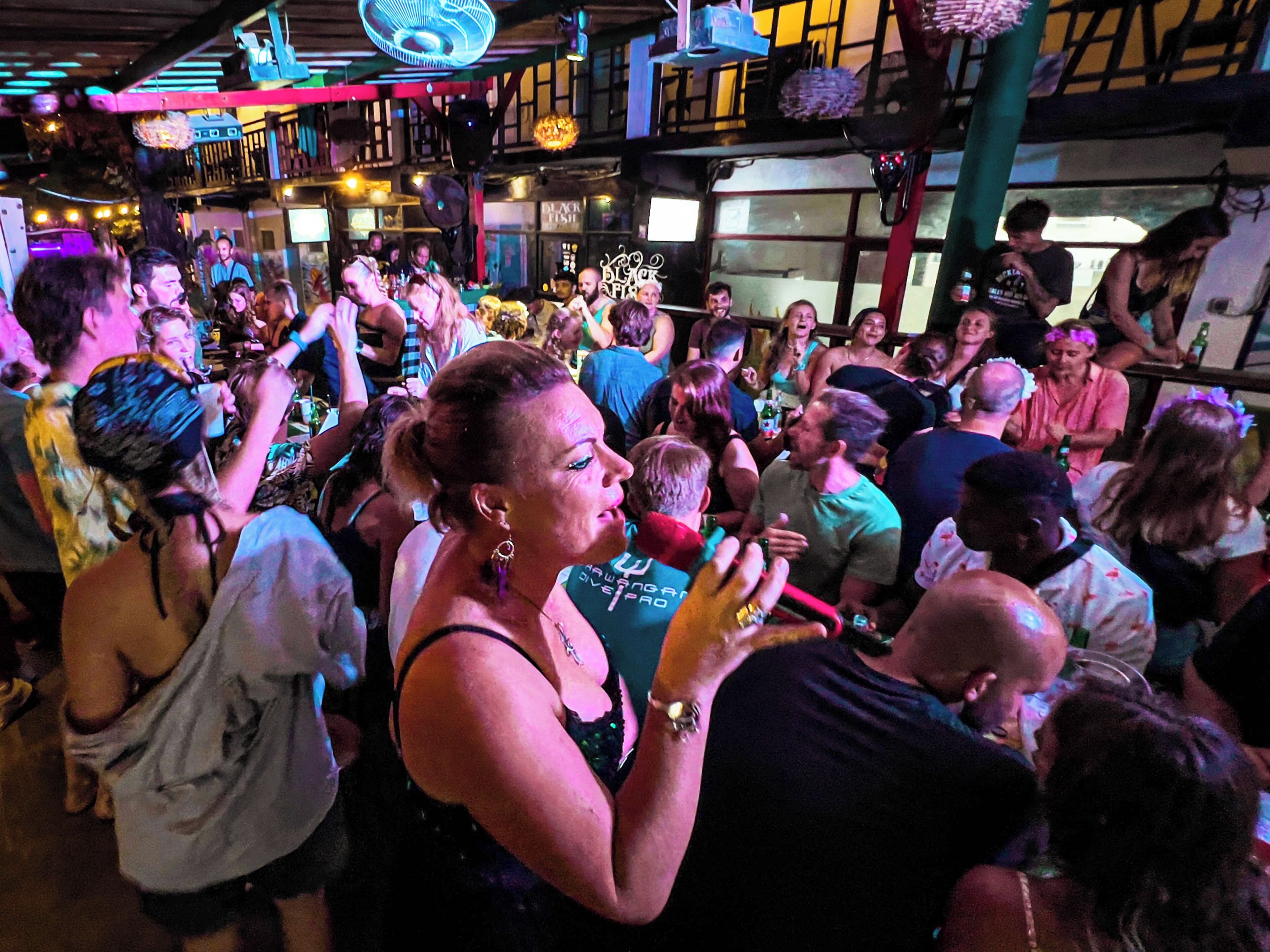 A crowded indoor venue with people gathered around tables, some holding drinks.