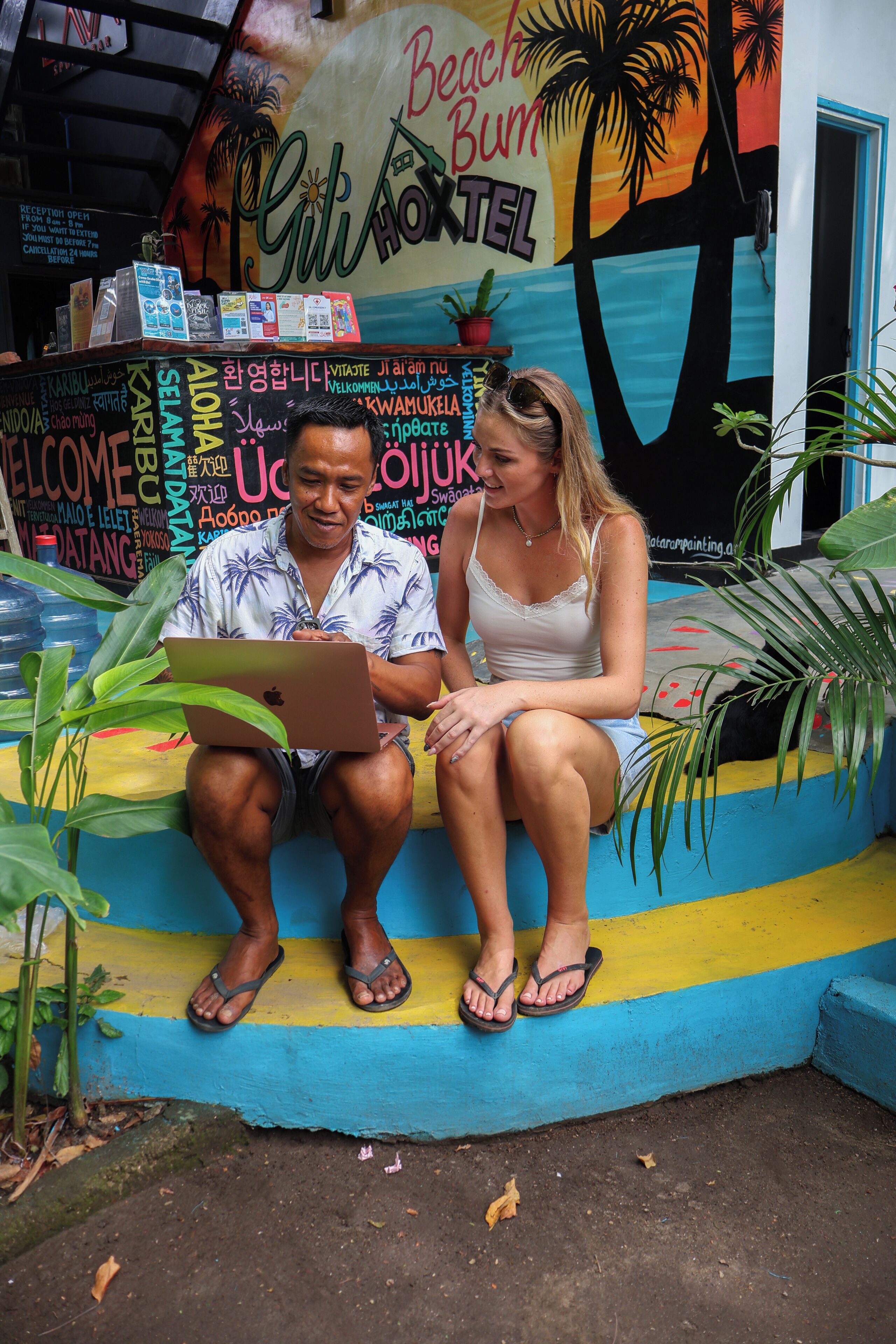 Dos personas sentadas en un banco colorido con un mural tropical al fondo.