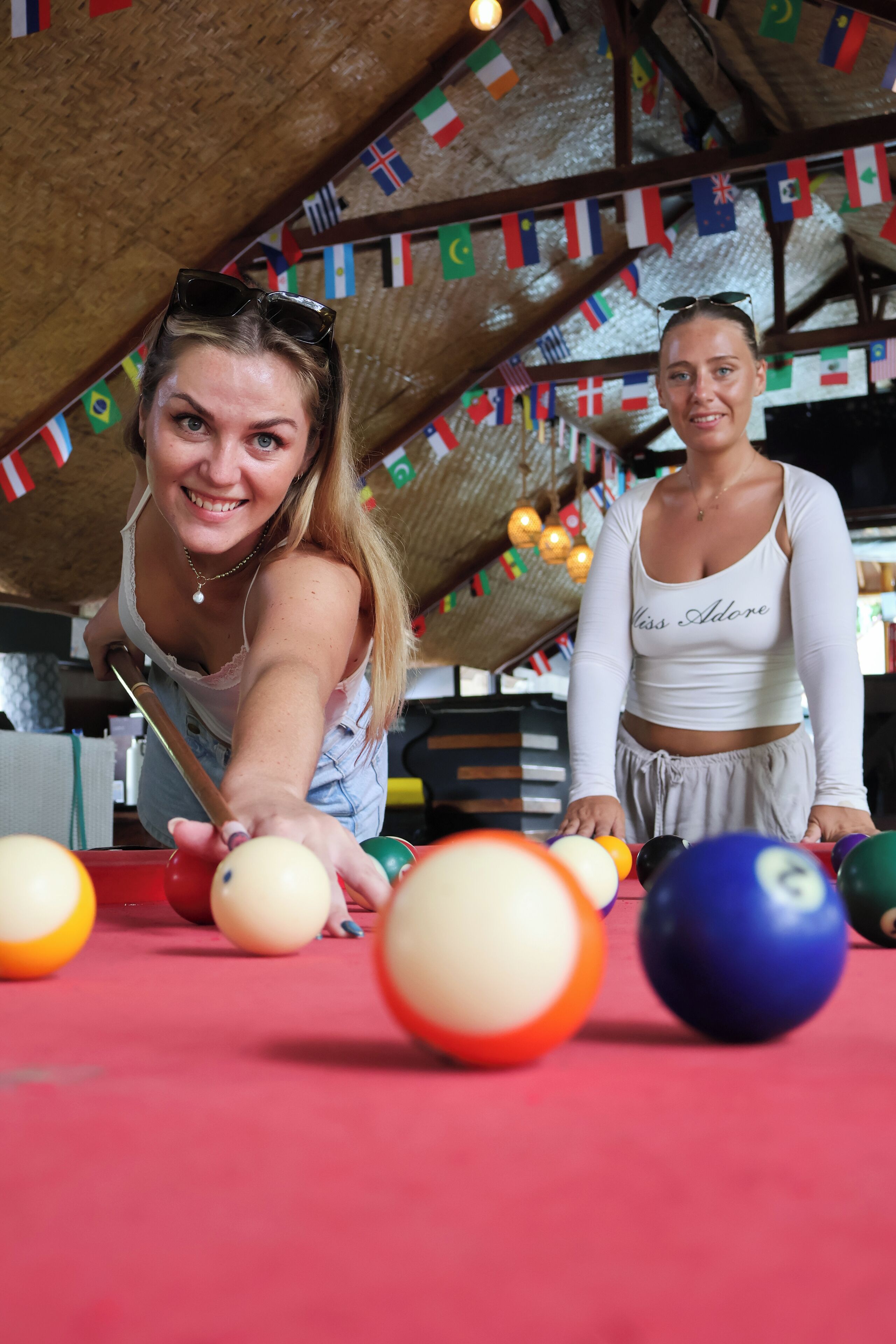 Two people playing pool in a room with a wooden ceiling and colorful flags.