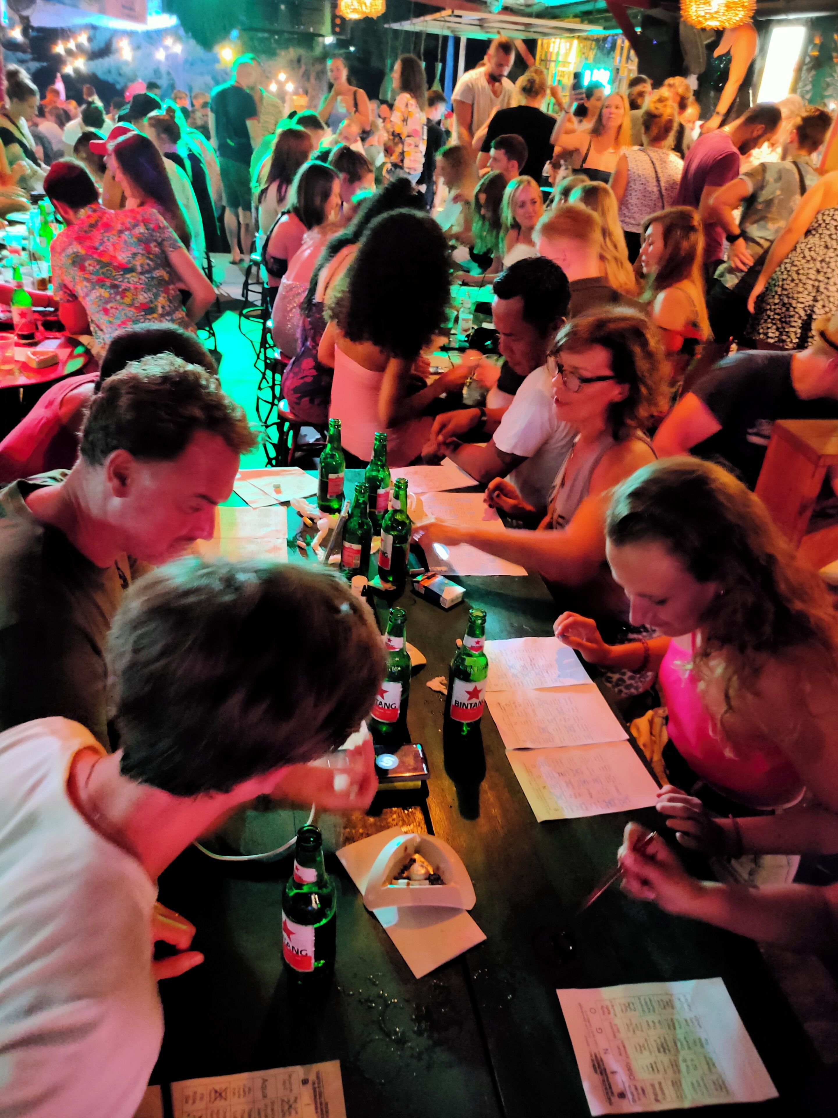 Eine belebte Bar mit an Tischen sitzenden Personen, einige mit Papieren und Flaschen.