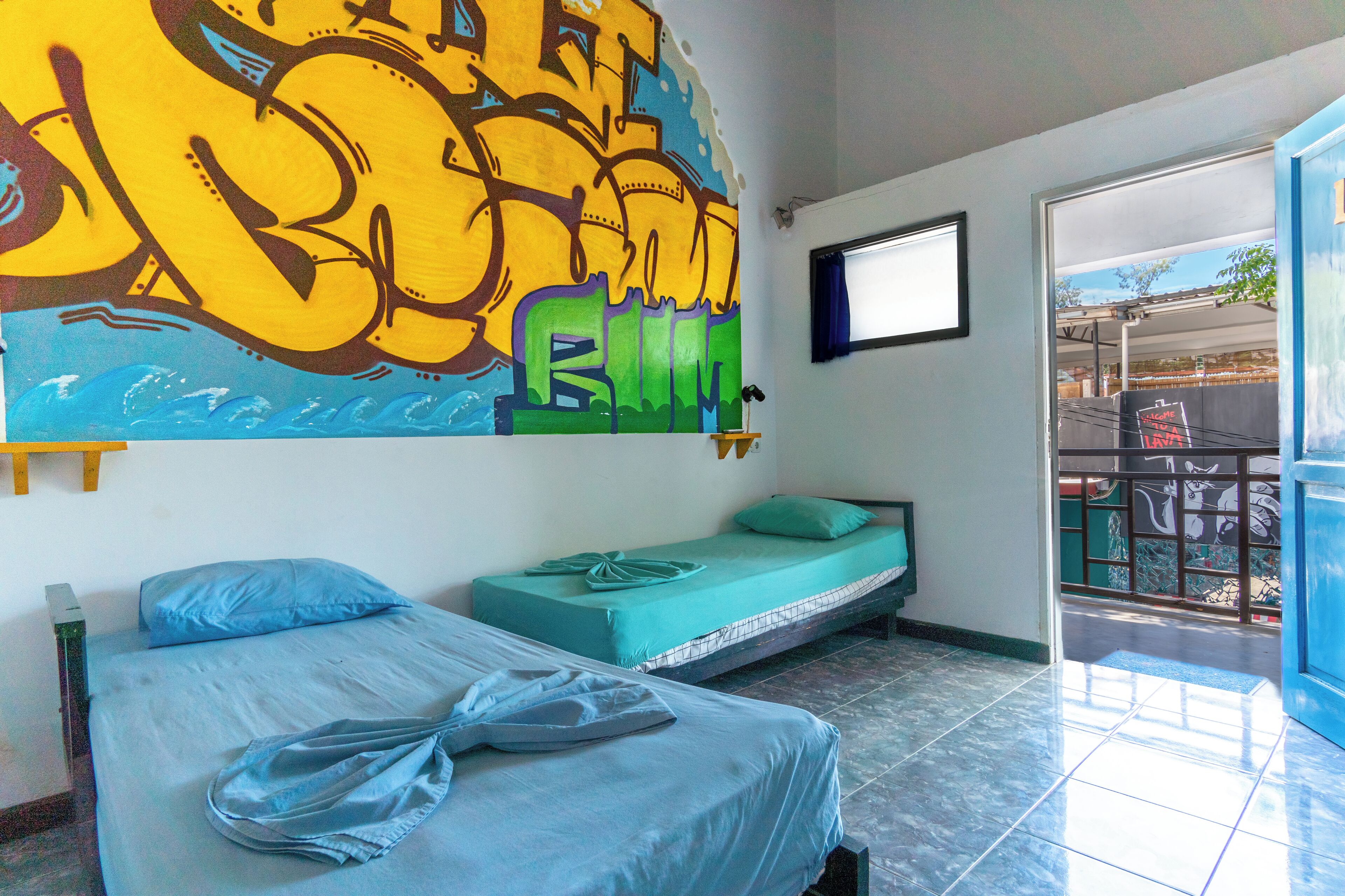 A room with two beds, one with a blue mattress and the other with a green mattress, a large graffiti artwork on the wall, and a window with a view of the outside.