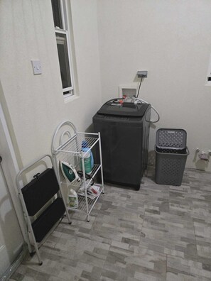 A small laundry area with a washing machine, a drying rack, a basket, and a radiator. - Cozy 4-bedroom bungalow in Brevitor, St. Peter, where you can live like a local. (Mile and A Quarter)
