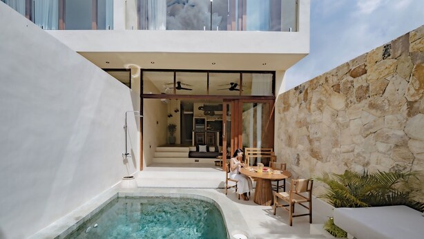 Un área de piscina moderna al aire libre con una pared de piedra, una zona de estar con mesa y sillas, y vistas hacia la casa.