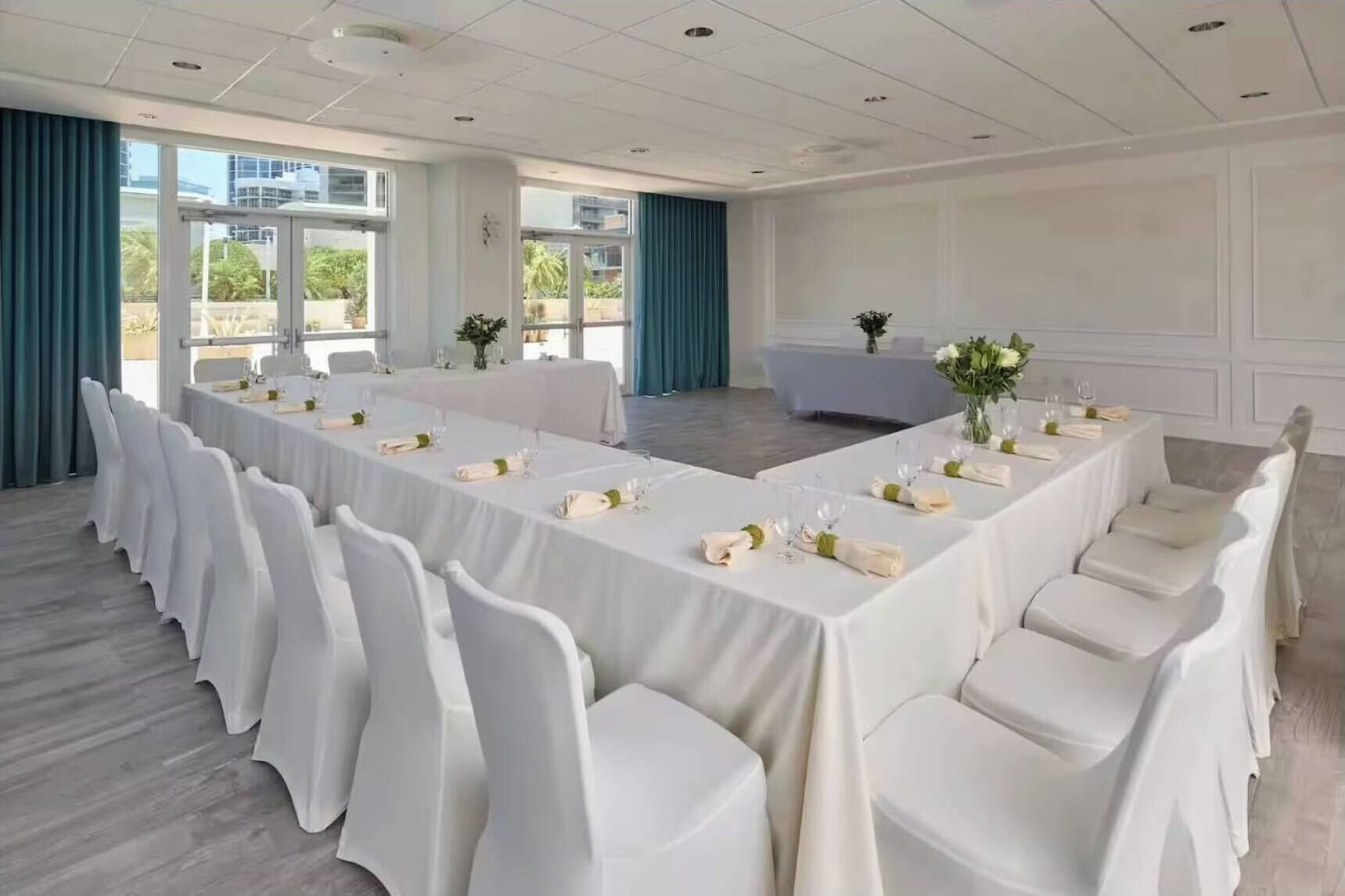Un salón de conferencias con una larga mesa dispuesta para una reunión, sillas blancas y un gran ventanal con vistas a edificios y zonas verdes.