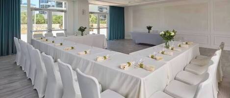 Un salón de conferencias con una larga mesa dispuesta para una reunión, sillas blancas y un gran ventanal con vistas a edificios y zonas verdes.
