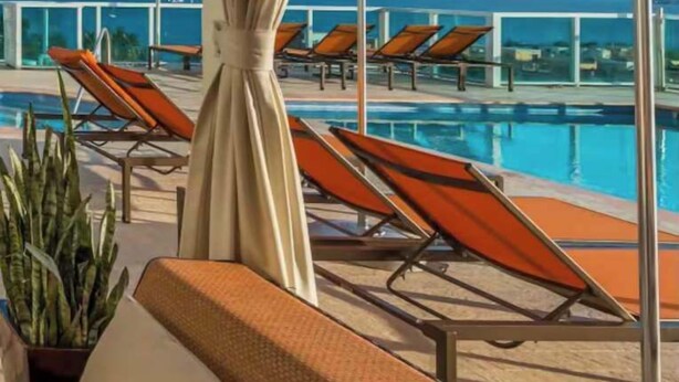 Zona de piscina con sillas de descanso y sombrillas, con vistas a un cielo azul despejado y una lejana vista del mar.