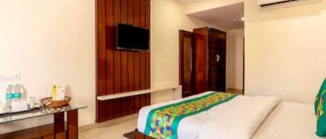 A hotel room with a bed, a TV mounted on the wall, a desk with a chair, and an air conditioning unit.
