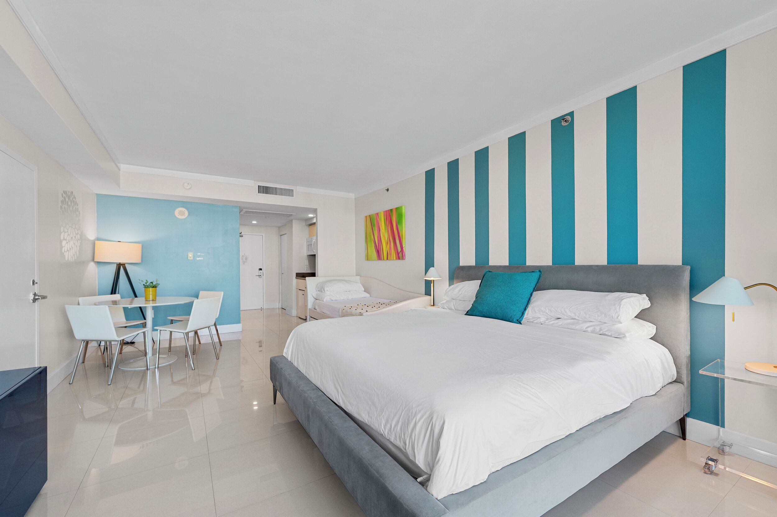 A modern hotel room with a large bed, a dining area with a table and chairs, and a bathroom visible through an open door.