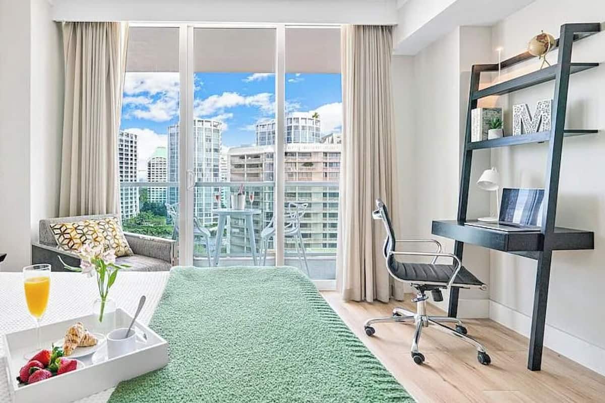 Una habitación de hotel moderna con un ventanal grande, una alfombra verde, un área de comedor con desayuno y un escritorio con una silla.
