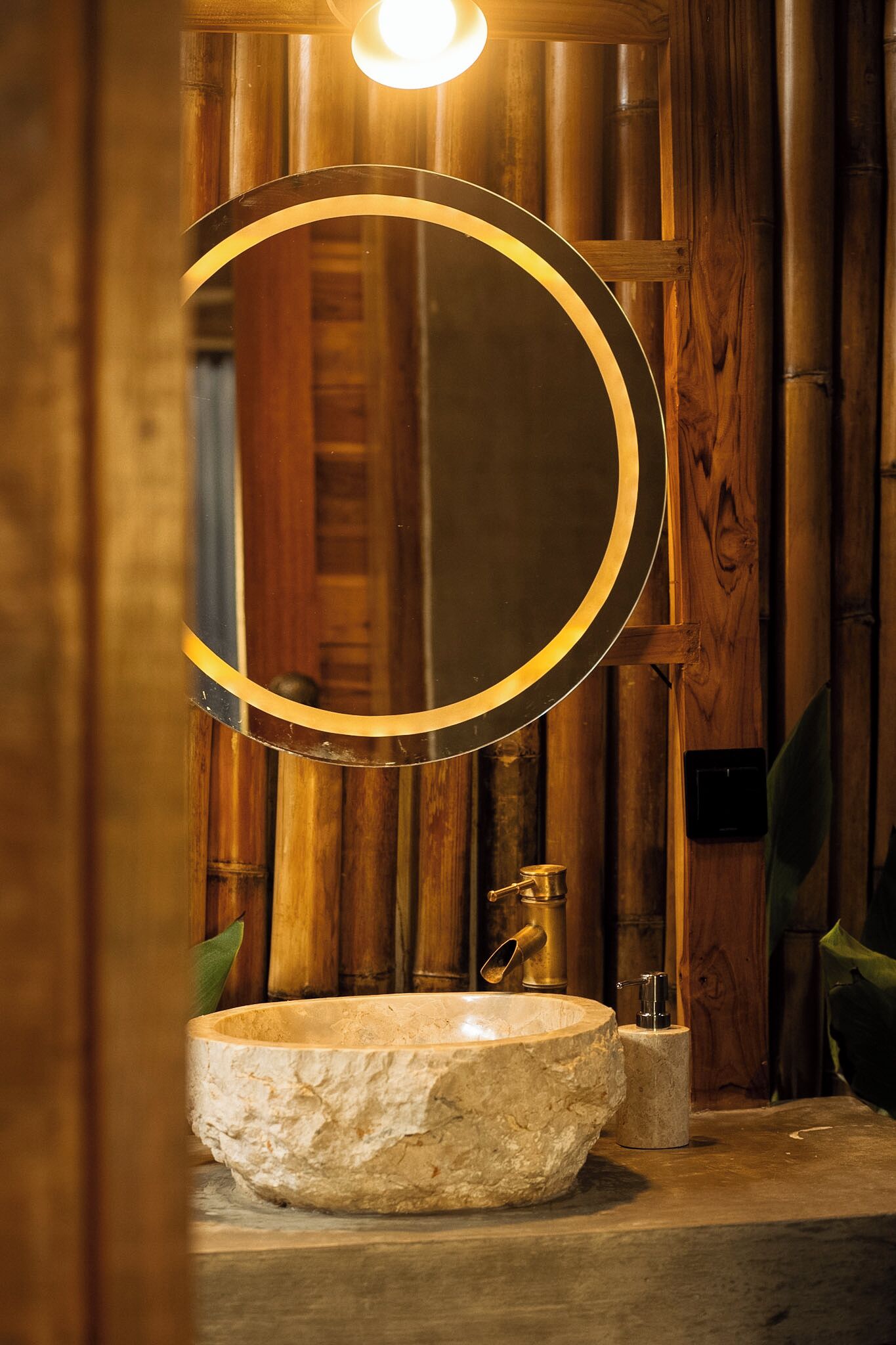A stone sink with a round mirror above it, set against a wooden wall with a bamboo backdrop.