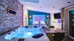 A modern room with a stone wall, a large bathtub, a bed with a blue wall, and a dining area with a table and chairs. - Loft Mini-Toy - Playful & Romantic Love Room (CARSPACH)
