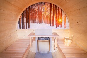 Una sauna con paredes de madera, un banco, un cubo y vistas a un bosque nevado a través de un ventanal arqueado.